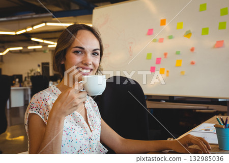 Professional woman sipping coffee from mug and smiling at office desk with whiteboard and pencils 132985036