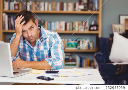 Man working on financial documents at home office desk with laptop calculator smartphone and papers Man working on financial documents at home office desk with laptop calculator smartphone and papers 132985039