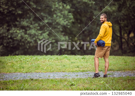 Man standing in clearing at forest edge holding rolled camping mat in mustard hoodie, copy space Man standing in clearing at forest edge holding rolled camping mat in mustard hoodie, copy space 132985040