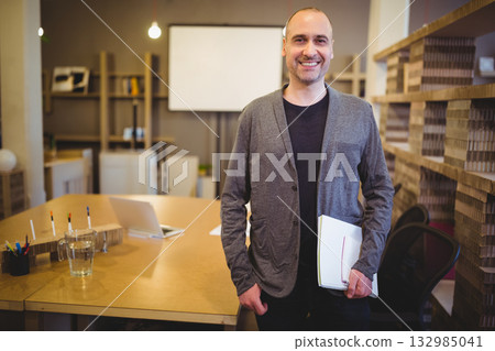 Man standing and smiling in modern office meeting area holding papers and notebook near laptop Man standing and smiling in modern office meeting area holding papers and notebook near laptop 132985041