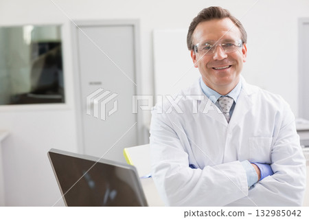 Middle-aged man in lab coat crossing arms at workbench with laptop and safety glasses, copy space 132985042