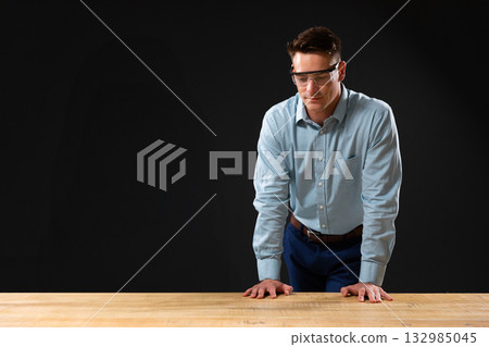 Male technician leaning on wooden worktable in studio workspace wearing safety glasses, copy space 132985045