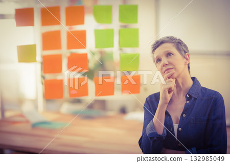 Senior woman standing at glass board with orange, green sticky notes in office, copy space 132985049