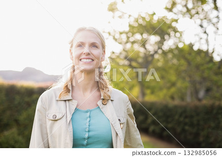Woman in her 30s smiling and looking upward while standing in sunlit garden wearing beige jacket Woman in her 30s smiling and looking upward while standing in sunlit garden wearing beige jacket 132985056