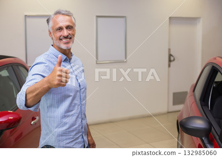 Man in his 50s giving thumbs-up and standing between red cars in dealership showroom 132985060