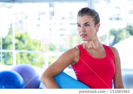 Woman wearing red top holding blue yoga mat in fitness studio with stability balls, copy space 132985062