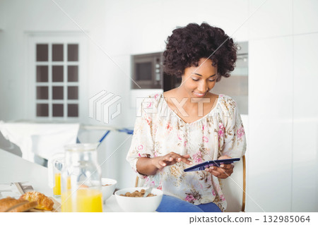 African American woman sitting at kitchen table using tablet with cereal bowl and juice, copy space African American woman sitting at kitchen table using tablet with cereal bowl and juice, copy space 132985064