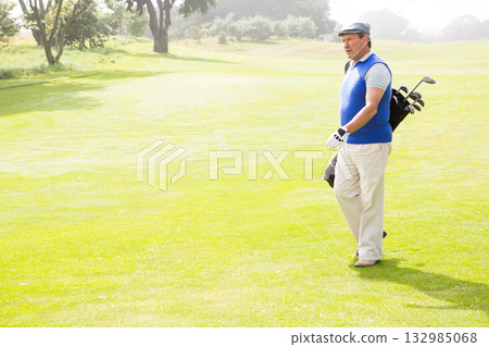 Senior African American man walking across fairway in golf attire carrying clubs in bag, copy space 132985068
