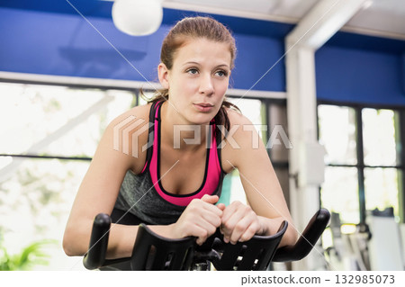 Female fitness enthusiast pedaling on stationary bike in gym wearing grey tank top and leggings 132985073