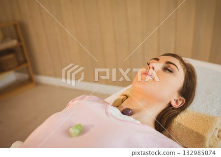 Female client lying on padded table at spa, receiving green chest, purple neck crystal therapy 132985074