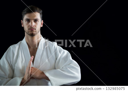 Male martial artist pressing hands at chest in studio with black backdrop, white gi, copy space 132985075