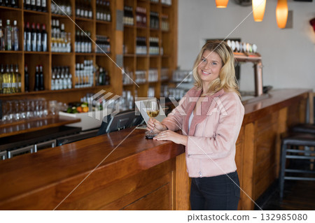 White female standing at bar counter holding wine glass under lights by bottle shelves, copy space 132985080