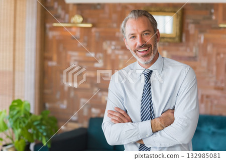 Businessman in shirt and tie standing arms folded in lounge with wood panels and teal sofa 132985081