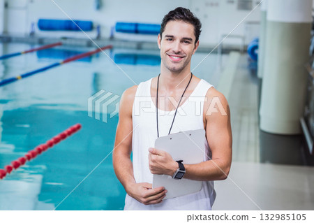 Male swim coach in tank top standing poolside holding clipboard wearing whistle near lane dividers 132985105