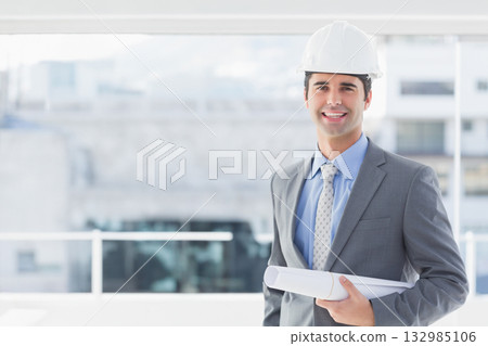 Male architect in suit standing on terrace overlooking city holding helmet and plans, copy space 132985106