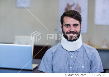 Male patient using silver laptop at medical clinic desk wearing cervical collar near anatomy charts Male patient using silver laptop at medical clinic desk wearing cervical collar near anatomy charts 132985108