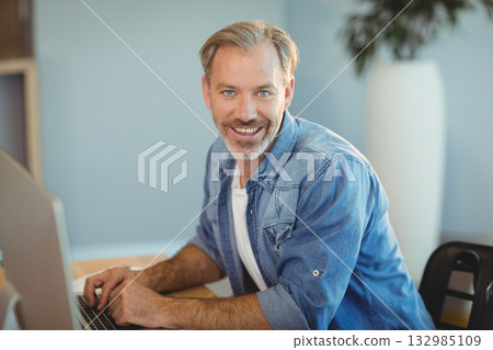 Middle-aged man typing on computer keyboard at wooden desk in bright office with potted plant 132985109