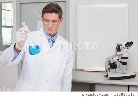 Male lab researcher holding conical flask with blue liquid at bench near microscope, copy space Male lab researcher holding conical flask with blue liquid at bench near microscope, copy space 132985112