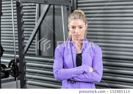 Woman standing with arms crossed in gym front of steel rack with weight plates wearing stopwatch 132985113