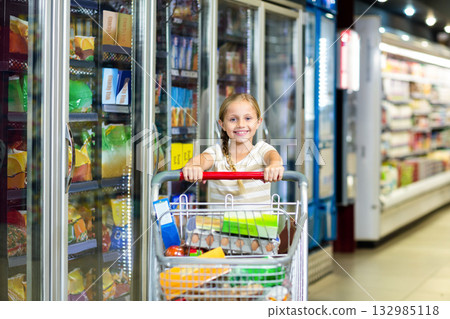 Girl pushing shopping cart loaded with eggs, cereal box and beverage cartons along grocery aisle 132985118
