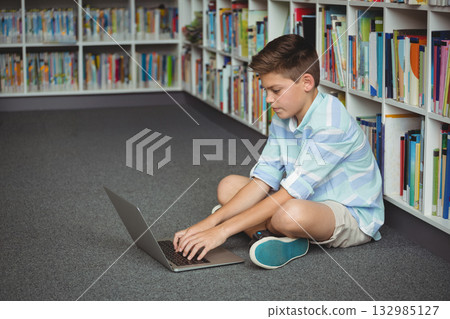 School-age boy sitting cross-legged in library using laptop near rows of bookshelves, copy space School-age boy sitting cross-legged in library using laptop near rows of bookshelves, copy space 132985127