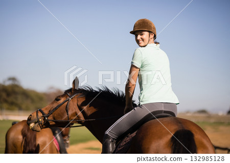 Female equestrian riding brown horse in sunny paddock wearing helmet and holding reins Female equestrian riding brown horse in sunny paddock wearing helmet and holding reins 132985128