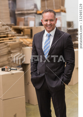 Middle-aged man wearing business suit standing in warehouse among boxes, pallets and tape dispenser Middle-aged man wearing business suit standing in warehouse among boxes, pallets and tape dispenser 132985129