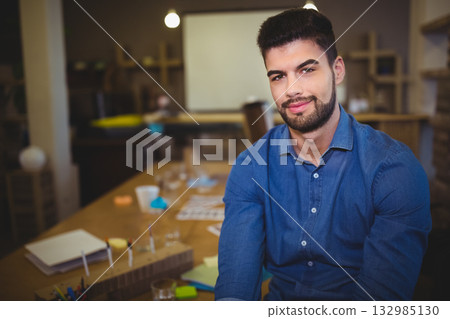 Male in his twenties sitting at table in office reviewing papers by pencil organizer, copy space 132985130