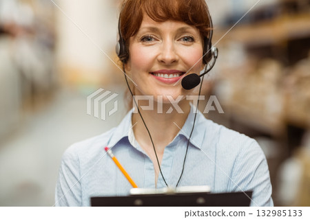 Woman standing in warehouse aisle wearing headset holding clipboard near metal shelving and boxes Woman standing in warehouse aisle wearing headset holding clipboard near metal shelving and boxes 132985133