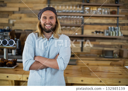Mid adult male barista smiling, crossing arms behind counter at coffee shop by espresso machine 132985139