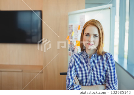 Adult female posing in checked shirt with arms crossed in meeting space by sticky-noted whiteboard Adult female posing in checked shirt with arms crossed in meeting space by sticky-noted whiteboard 132985143