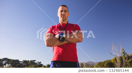 Male fitness coach standing with arms crossed in sportswear on field with stopwatch, wrist wrap 132985144