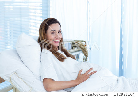 Pregnant woman reclining on hospital bed wearing hospital gown, holding abdomen with monitor nearby 132985164