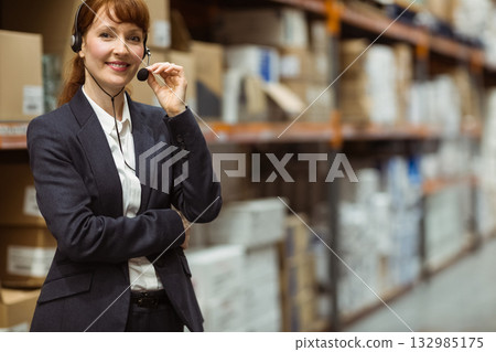 Mid adult supervisor touching headset mic in warehouse aisle by boxes on metal shelves, copy space 132985175