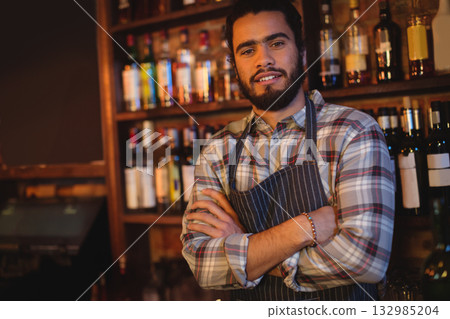 Male bartender wearing apron standing behind bar counter with arms crossed amid liquor bottles 132985204