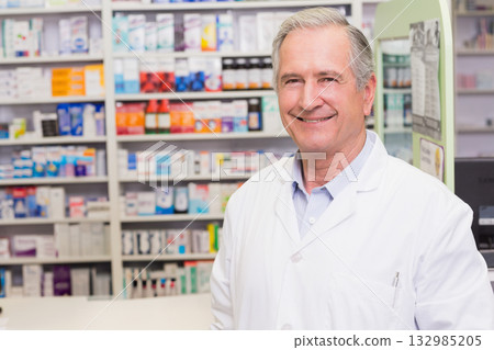 Senior male pharmacist standing behind pharmacy counter by medicine shelves, info board, copy space 132985205