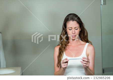 Woman holding pregnancy test stick at chest, examining result with concern in bathroom, copy space Woman holding pregnancy test stick at chest, examining result with concern in bathroom, copy space 132985206