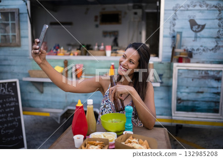 Woman taking selfie at wooden table in front of food truck holding beer bottle and smartphone 132985208