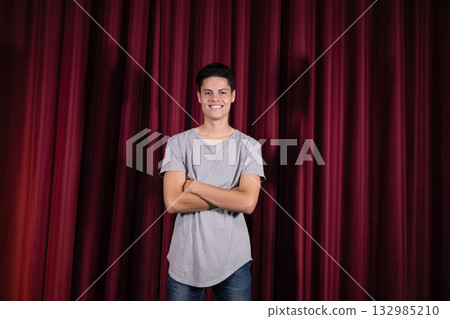 Hispanic man standing with arms crossed on stage wearing tee and jeans before maroon-red curtains Hispanic man standing with arms crossed on stage wearing tee and jeans before maroon-red curtains 132985210