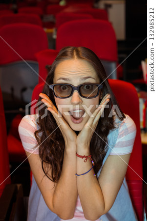Female moviegoer wearing viewing glasses sitting in red cinema seats leaning forward in surprise 132985212