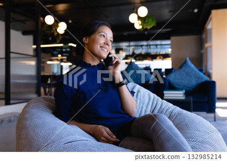 Asian woman sitting in office lounge holding smartphone to ear beside blue sofa with pendant lights Asian woman sitting in office lounge holding smartphone to ear beside blue sofa with pendant lights 132985214