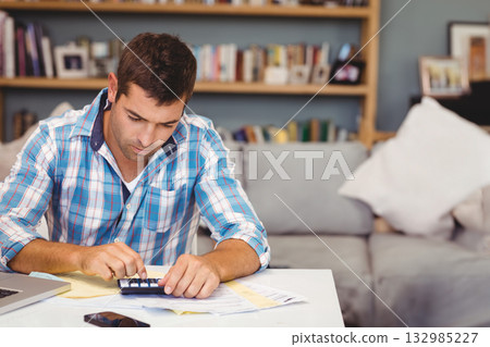 Man sitting at table in living room using calculator with laptop phone and documents, copy space Man sitting at table in living room using calculator with laptop phone and documents, copy space 132985227