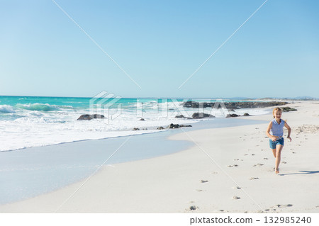 Female child running barefoot along sandy shore beside turquoise waves and dark rocks, copy space 132985240