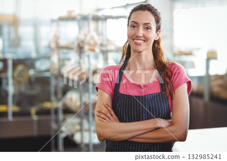 Female baker standing behind counter in bakery wearing striped apron and smiling with bread shelves 132985241