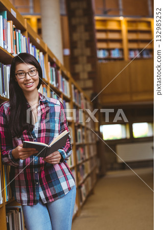 Asian woman wearing eyeglasses leaning against bookshelf in library holding open book and smiling 132985252