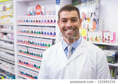 Male pharmacist standing in pharmacy aisle with medicine boxes wearing coat and tie, copy space 132985259