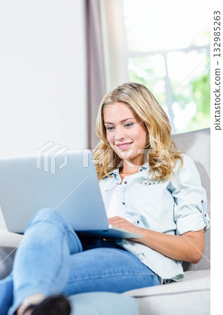 Woman sitting on light gray sofa near window, typing on silver laptop while studying remotely 132985263