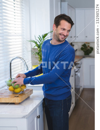 Man wearing blue sweater placing apples and lemons into glass bowl on cutting board in kitchen Man wearing blue sweater placing apples and lemons into glass bowl on cutting board in kitchen 132985274