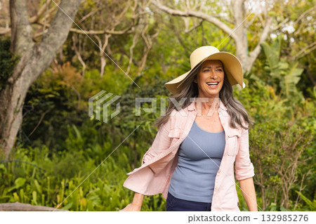 Senior woman walking along garden path wearing wide-brimmed straw sun hat and casual clothing Senior woman walking along garden path wearing wide-brimmed straw sun hat and casual clothing 132985276