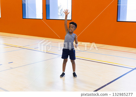 African American boy raising right arm while standing on multi-sport court under tall windows African American boy raising right arm while standing on multi-sport court under tall windows 132985277